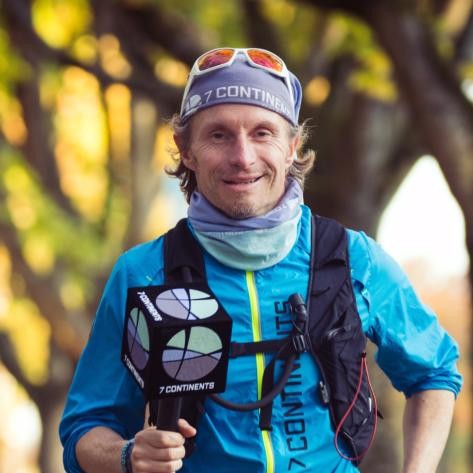 Norman Bücher - 7 CONTINENTS - in 20.000 km um die Welt (Multivisionsvortrag) Norman Bücher steht im Freien und lächelt in die Kamera. Er trägt eine blaue Outdoor-Jacke, ein multifunktionales Halstuch, eine Sonnenbrille auf dem Kopf